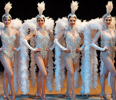 Four female performers in silver and white costumes with feather collars